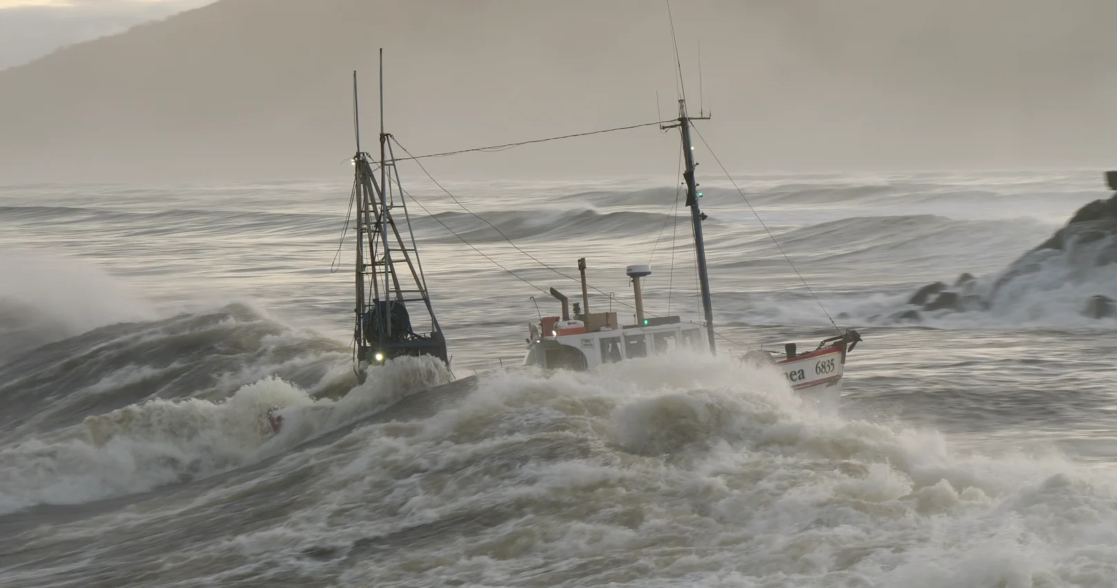 Fishing vessel crossing the Greymouth bar in rough seas — waves crashing, masts cutting through the swell